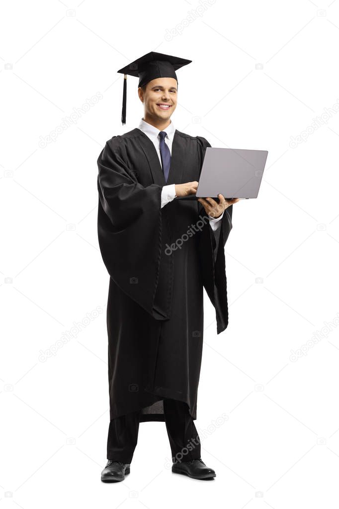 Full length portrait of a young man in a black graduation gown and cap holding a laptop computer and smiling at the camera isolated on white background