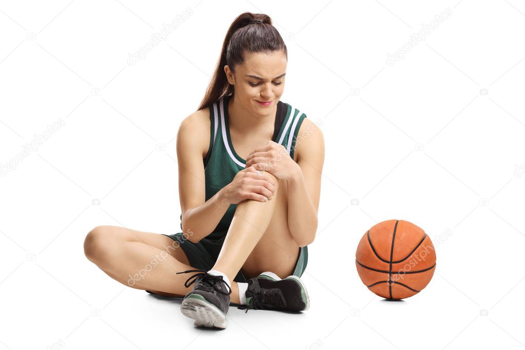 Young female basketball player sitting on a floor and holding her painful knee isolated on white background