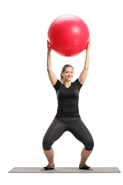 一个年轻女性在白色背景上隔离的运动垫上举起健身球的全长肖像 — 图库照片