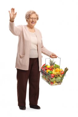 Eliyle beyaz arka plan üzerinde izole el sallayarak bir alışveriş sepeti meyve ve sebze ile yaşlı bir kadının tam uzunlukta portre