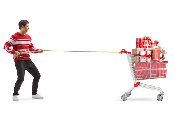 白い背景に孤立したロープでプレゼントでいっぱいのショッピングカートを引っ張る若者のフルレングスショット — ストック写真