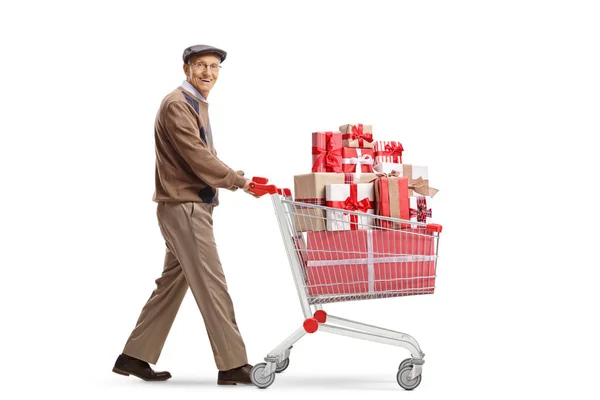 白い背景に隔離されたカメラに微笑むプレゼントでいっぱいのショッピングカートを持つ高齢者のフルレングスショット — ストック写真