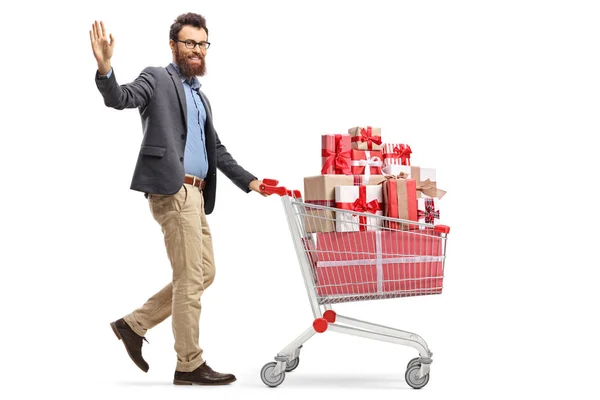 白い背景に隔離されたプレゼントでいっぱいのショッピングカートで手を振って歩いているあごひげの男のフルレングスショット — ストック写真