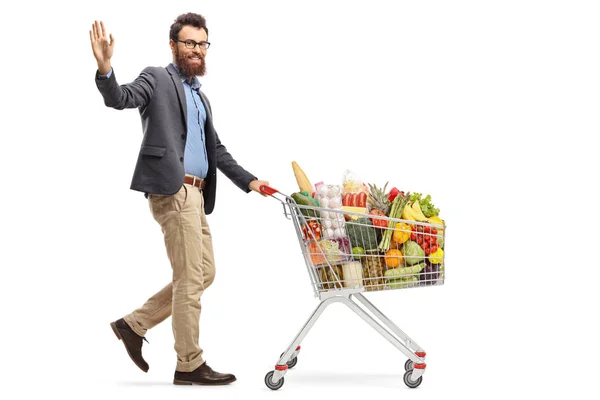 食料品を持つショッピングカートを持って歩き 白い背景に孤立した手で手を振る男のフルレングスショット — ストック写真