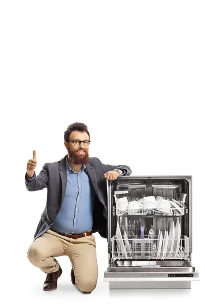 ひげを生やした男が食器洗い機の隣にひざまずき 白い背景に孤立した親指を見せる — ストック写真