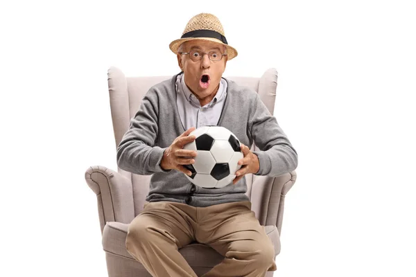 Ενθουσιασμένος ηλικιωμένος άνθρωπος κρατώντας ένα ποδόσφαιρο και κάθεται σε ένα βραχίονα — Φωτογραφία Αρχείου