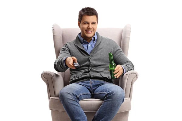 白い背景に隔離された肘掛け椅子のリモコンとビール瓶を持つ若い男 — ストック写真