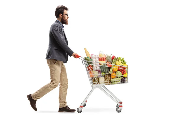 白い背景に隔離された食品でショッピングカートを歩いて押すあごひげの男のフルレングスショット — ストック写真