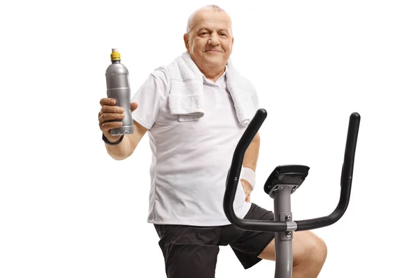 Ώριμη άνθρωπος σε ένα ποδήλατο άσκησης κρατώντας ένα μπουκάλι νερό — Φωτογραφία Αρχείου