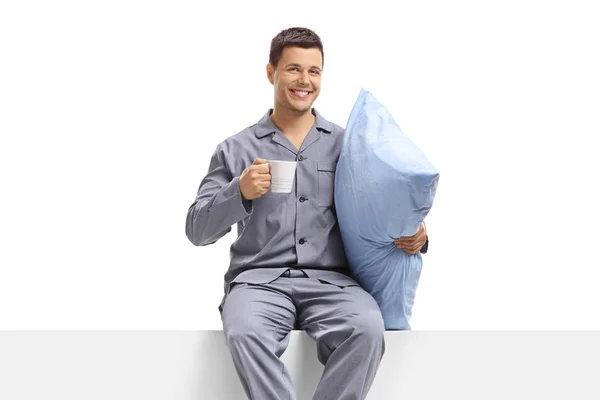 Mann im Pyjama hält eine Tasse und ein Kissen in der Hand und sitzt auf einer Platte — Stockfoto