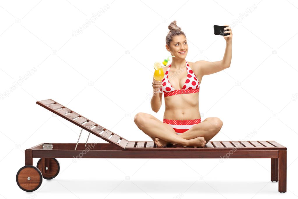 Young female in bikini drinking a cocktail and taking a selfie w