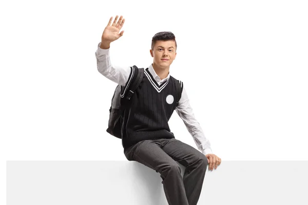 Estudante adolescente masculino em um uniforme sentado em um painel e acenando — Fotografia de Stock