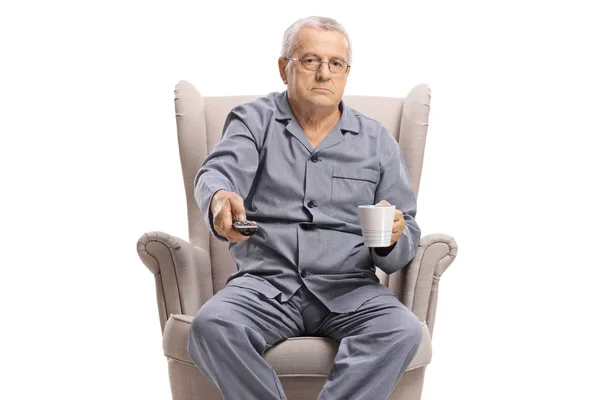 Elderly man holding a remote control and a cup sitting in an arm — Stock Photo, Image