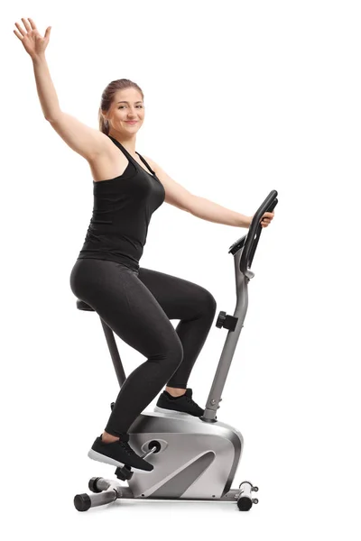 Young female riding a stationary bike and waving at camera — Stok fotoğraf