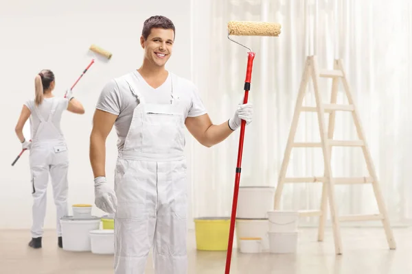 Male painter with a paint roller posing and a female painter painting a wall inside a house