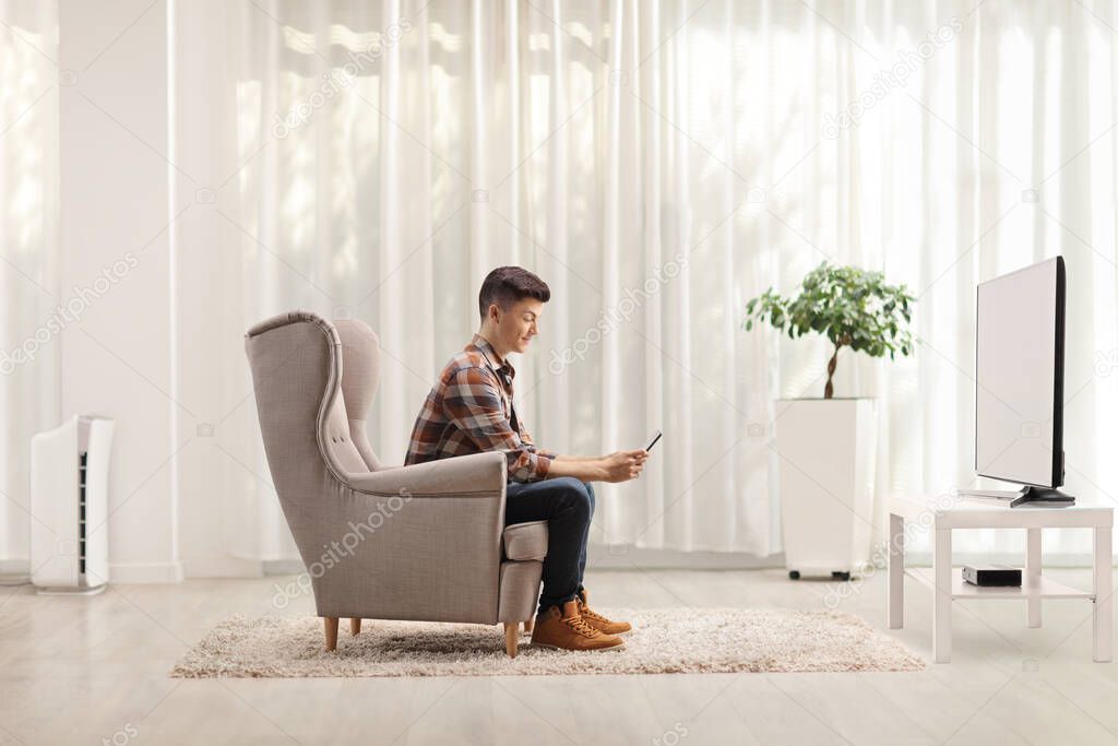 Guy sitting at home in an armchair and using a mobile phone in front of a tv