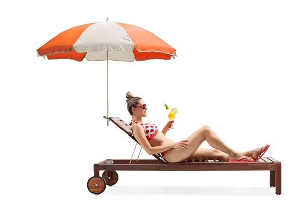 一个身穿比基尼的年轻女子躺在日光浴床上 拿着雨伞 喝着与白色背景隔离的鸡尾酒 照片全长拍摄 — 图库照片