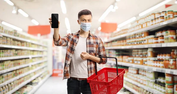 保護顔のマスクをしたスーパーマーケットの男と携帯電話を示すショッピングバスケット — ストック写真