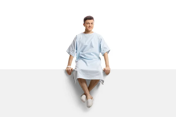 身穿医院长袍的年轻男性患者坐在面板上 凝视着白色背景的相机 — 图库照片