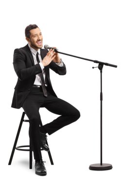 Bir sandalyede oturan ve beyaz arka planda izole edilmiş bir mikrofonda şarkı söyleyen takım elbiseli bir erkek şarkıcı.