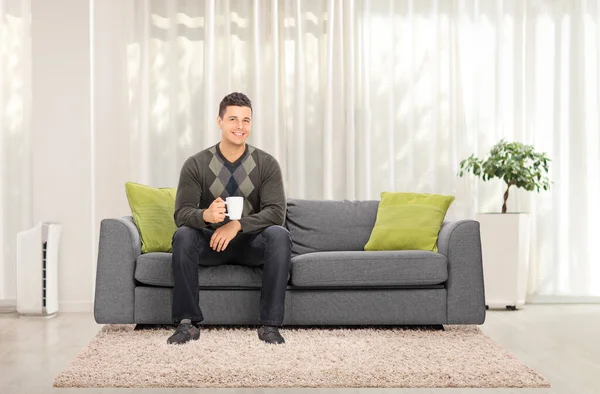 自宅でソファに座ってお茶を飲んでいるカジュアルな男の完全な長さの肖像画 — ストック写真