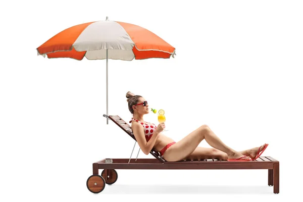 一个身穿比基尼的年轻女子躺在日光浴床上 在白色背景的雨伞下喝着鸡尾酒的全身像照片 — 图库照片
