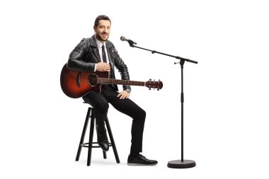 Beyaz arka planda mikrofonlu bir sandalyede oturan akustik gitarlı bir erkek şarkıcı.