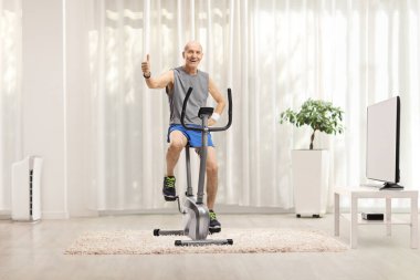 Spor kıyafetleri içinde neşeli yaşlı bir adam durgun bir bisiklette oturuyor ve evde baş parmağıyla işaret veriyor.