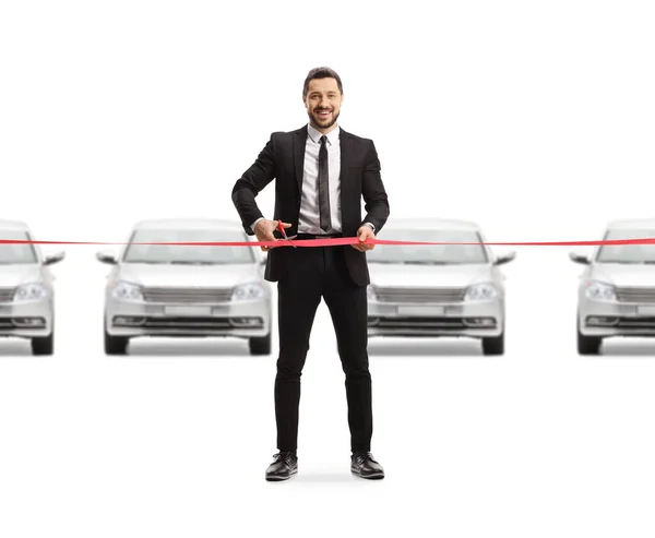 一个穿西装的男人的全长肖像 在汽车展示室里剪下红带带 对着被白色背景隔离的相机笑着 — 图库照片