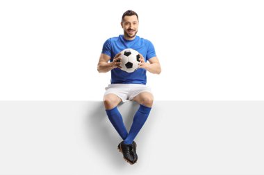 Futbolcu boş bir panelde oturuyor ve beyaz arka planda izole edilmiş bir futbol topu tutuyor.