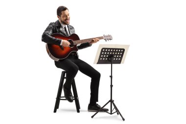 Akustik gitar çalan müzisyen ve beyaz arka planda izole edilmiş bir tezgahta duran bir müzik defterine bakıyor.