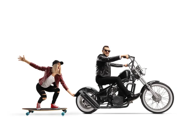 Full length profile shot of a biker riding a chopper and pulling a female on a skateboard isolated on white background