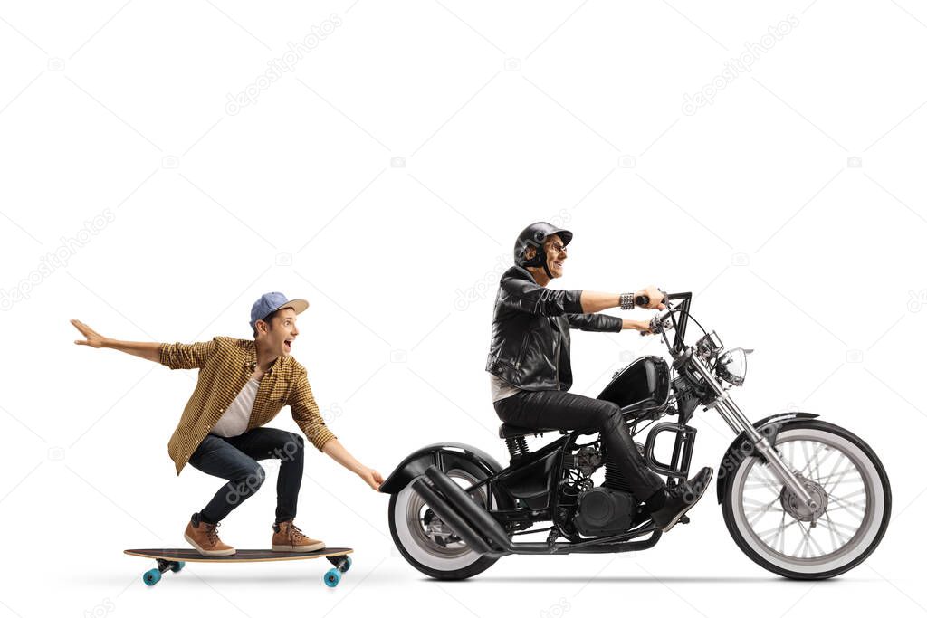 Full length profile shot of an elderly biker riding a chopper and pulling a guy on a skateboard isolated on white background