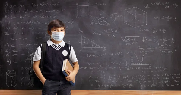 フェイスマスクを着用し 数式と学校の黒板の前に立って本やバックパックを持つ学校の少年 — ストック写真