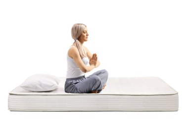 Pijamalı genç bir kadın beyaz arka planda izole edilmiş bir yatakta yoga meditasyonu yapıyor.