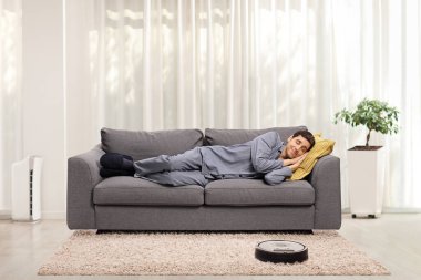 Robotik süpürge, pijamalı bir adam oturma odasında kanepede uyurken halıyı süpürüyor.