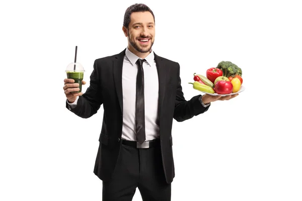 健康的な緑のスムージーと白い背景に隔離された果物や野菜のプレートを保持スーツで笑顔のプロの男 — ストック写真