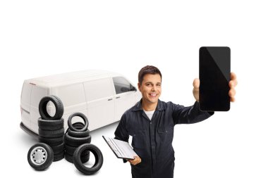 Beyaz minibüslü genç bir otomobil tamircisi elinde bir pano ve beyaz arka planda izole edilmiş bir akıllı telefon gösteriyor.