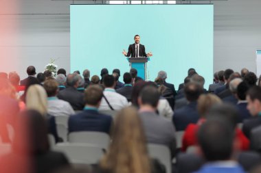 İnsanlar konferans salonunda oturuyor ve takım elbiseli bir adam konuşma yapıyor. 