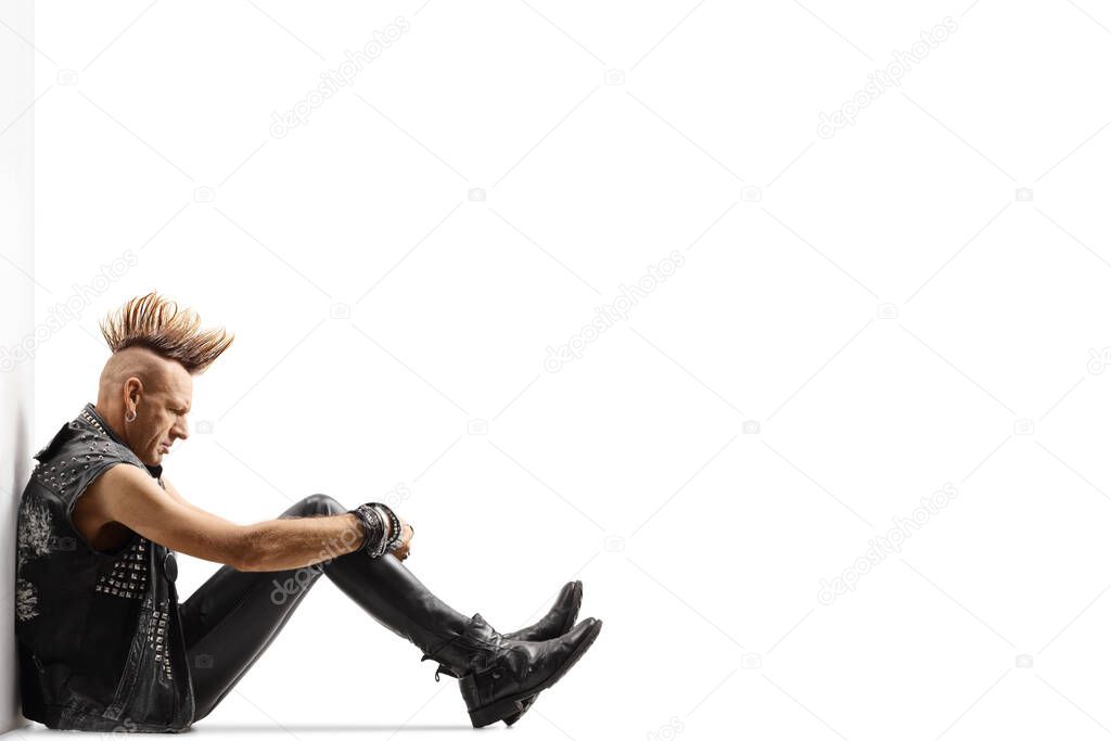 Pensive punk rocker with a mohawk sitting on the ground and leaning on a wall isolated on white background