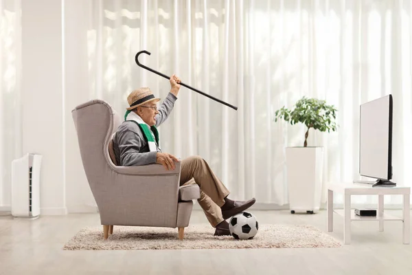 Elderly man soccer fan with cane and a scarf sitting in an armchair and cheering in front of tv at home