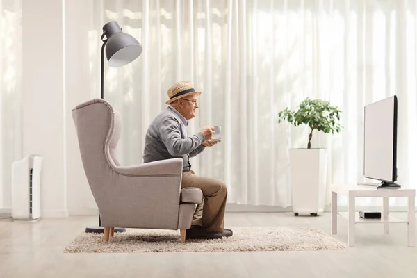 Elderly man with a cup sitting in an armchair and watching tv at home