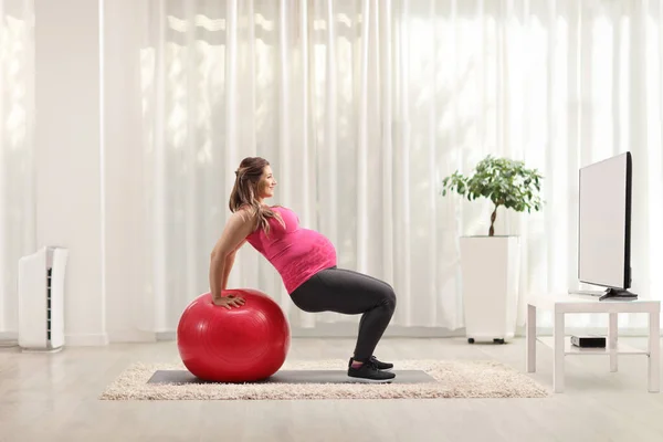 テレビの前でフィットネスボールを持って家で妊娠中の練習をしている女性 — ストック写真