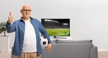 Olgun bir adam televizyon ekranının önünde, kanepe elinde futbol topu tutuyor ve baş parmağını kaldırıyor. 