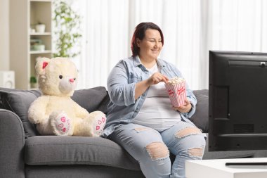 Kanepede bir oyuncak ayıyla oturan, televizyon izleyen ve patlamış mısır yiyen bir kadın.