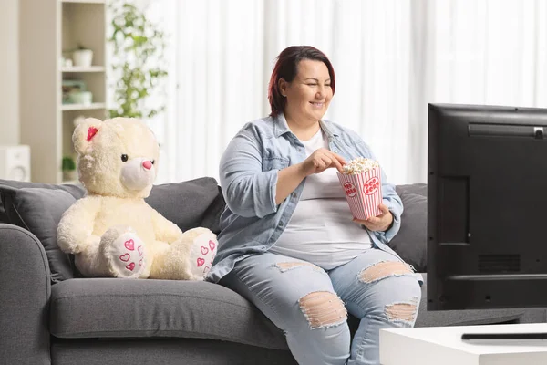 Kanepede bir oyuncak ayıyla oturan, televizyon izleyen ve patlamış mısır yiyen bir kadın.