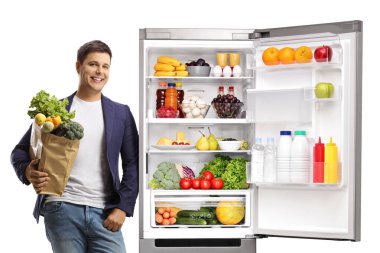 Beyaz arka planda izole edilmiş yiyeceklerle dolu açık bir buzdolabına yaslanmış genç bir adam.