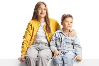 Genç kız kardeşi ve erkek kardeşi boş bir panelde oturuyor ve beyaz arka planda izole edilmiş kameraya gülümsüyor.