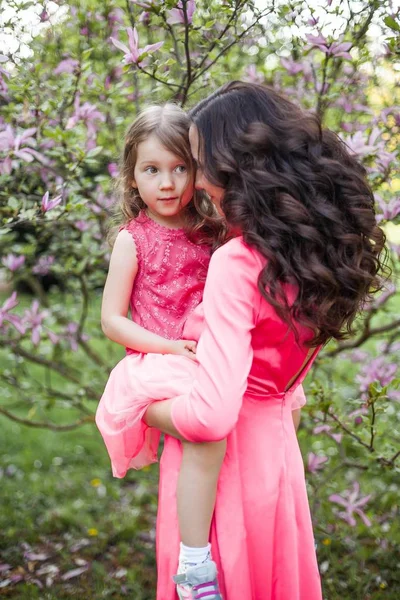 Uma jovem mãe bonita com uma filha pequena está andando ao longo do parque da primavera. Retrato de uma mãe e uma filha perto da magnólia florescente. Primavera. Uma família feliz. Calor . — Fotografia de Stock