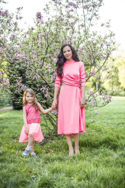 A young beautiful mother with a little daughter is walking along the spring park. Portrait of a mother and a little daughter near the blooming magnolia. Spring. A happy family. Heat. — Stock Photo, Image